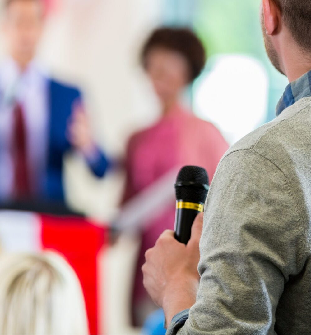 Image d'un homme de dos, un micro à la main afin de poser une question lors d'un meeting politique