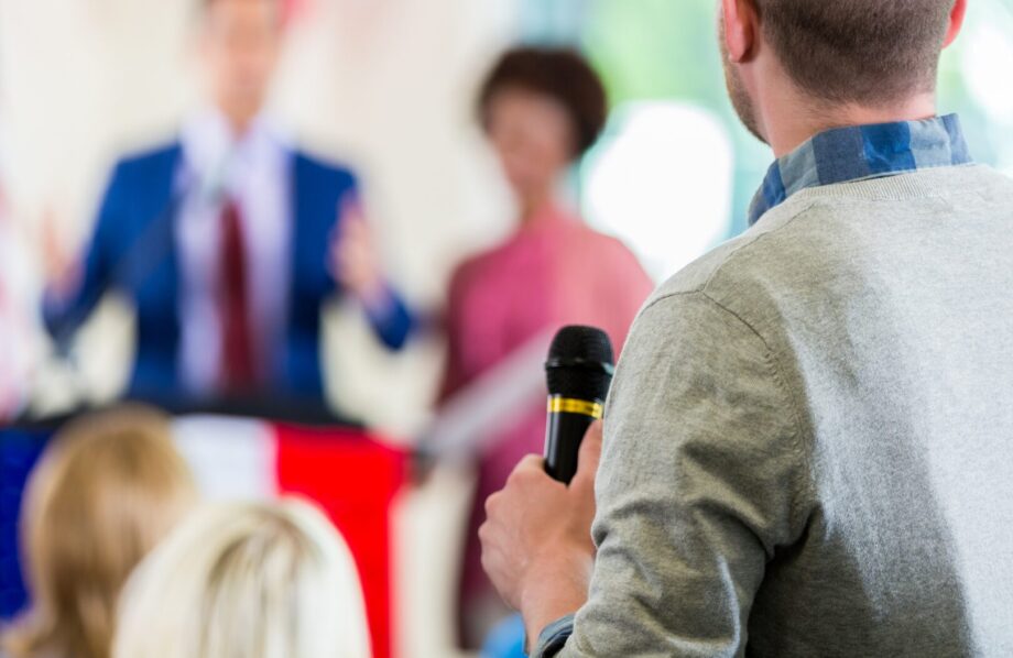 Image d'un homme de dos, un micro à la main afin de poser une question lors d'un meeting politique