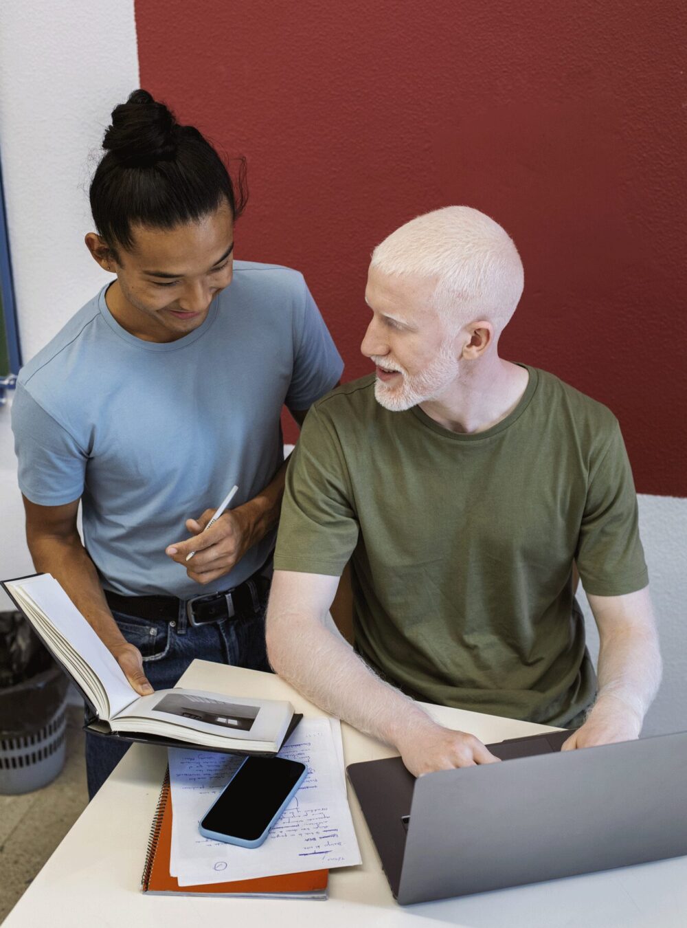Deux collègues qui échangent de manière informel. L'un écrit sur son ordinateur et est assis et l'autre est debout et lui montre un carnet