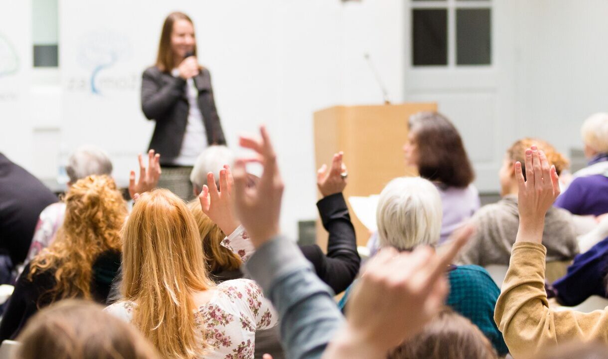 Image d'un conférence. Une femme prend la parole sur scène et au premier plan des mains sont levées