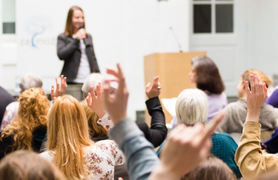 Image d'un conférence. Une femme prend la parole sur scène et au premier plan des mains sont levées